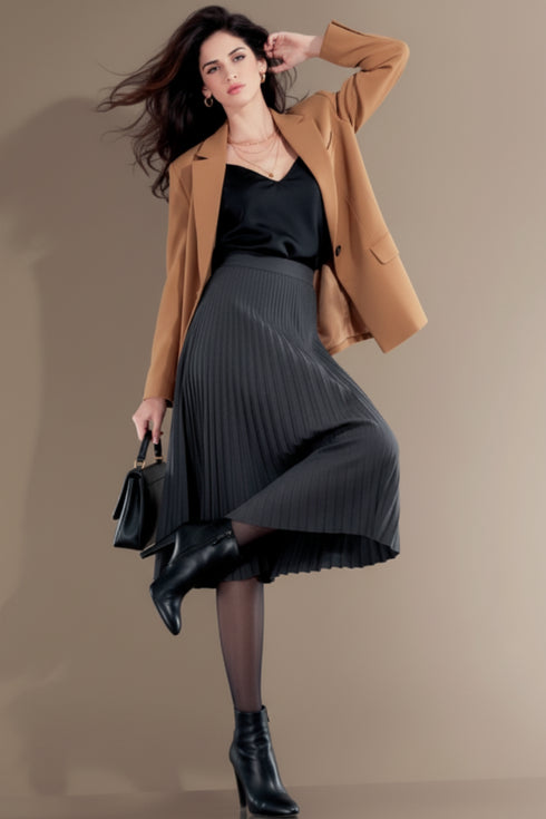 Woman wearing a brown blazer, black top, and gray pleated skirt with black shoes against a beige background.