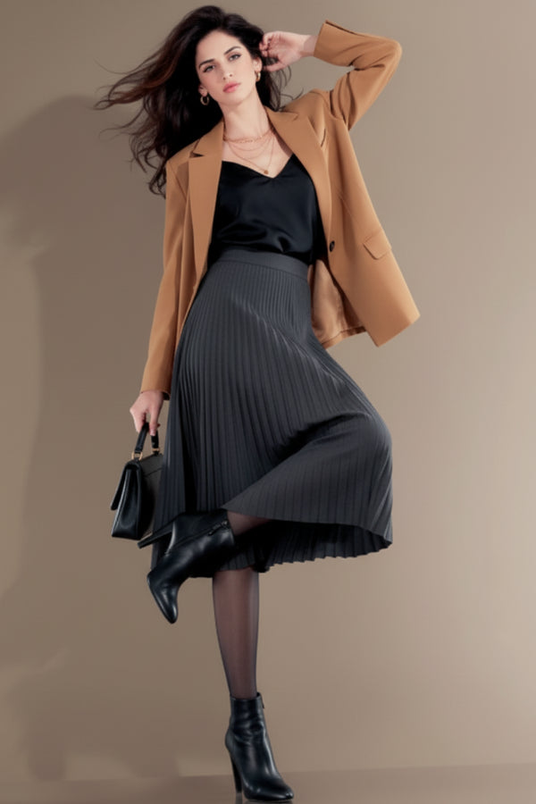 Woman wearing a brown blazer, black top, and gray pleated skirt with black shoes against a beige background.