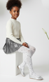 Child sitting on a stool wearing a white sweater, gray skirt, and white shoes with polka dots.