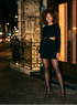 Woman in a black dress and stockings standing on a city street at night.