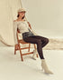 Woman sitting on a chair wearing a white off-shoulder top, denim shorts, black tights, and white high-heeled boots.