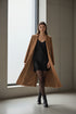 Full-body fashion editorial shot of a model wearing black 20 denier sheer tights, black silk slip dress, structured camel wool coat open at front, and black leather ankle boots. Minimal luxe setting with soft natural light. The pose is effortless—standing with coat draped elegantly, one leg slightly forward. Quiet luxury aesthetic: refined neutrals, elevated layering, timeless sophistication. Clean, high-end composition.