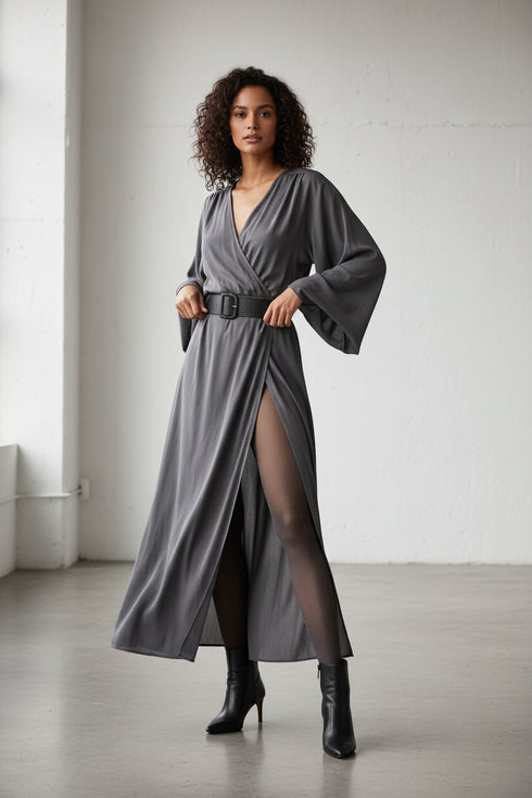 Full-body fashion editorial shot of a model wearing graphite 20 denier sheer-to-waist tights, graphite wrap dress cinched at waist with leather belt, and black leather ankle boots. Soft natural lighting in minimal setting. The pose is elegant and relaxed—one hand adjusting belt, confident stance. Emphasis on clean waistline with no visible seam line under the belt. Effortless sophistication, polished femininity, modern editorial diverse model