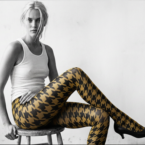 Woman wearing a white tank top and patterned tights sitting on a stool against a white background.