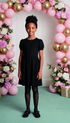 Young girl in a black dress standing in front of a decorative arch with balloons and flowers.