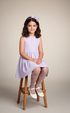 Young girl in a light purple dress sitting on a wooden stool against a plain background