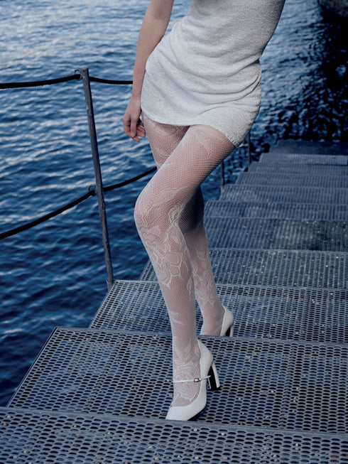 Person wearing a white dress and fishnet stockings standing on a dock by the water.