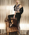 Corallo leggings feature a sleek black base with a floral lace pattern along the side, worn with a fitted top and layered jacket, seated on a brown leather chair amidst a textured backdrop.