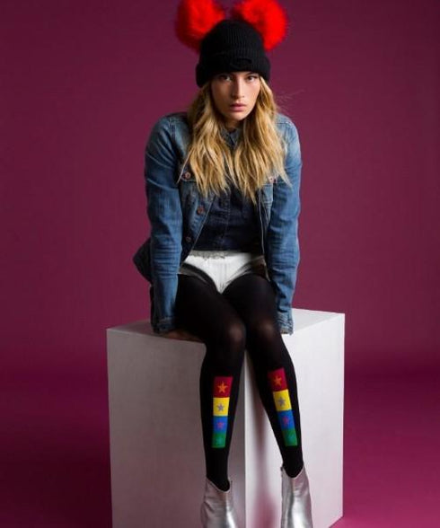 United Colors Opaque Tights feature a black base with a colorful pattern of rectangles and stars in red, yellow, and green, worn with a denim jacket, white shorts, and silver boots against a purple background.